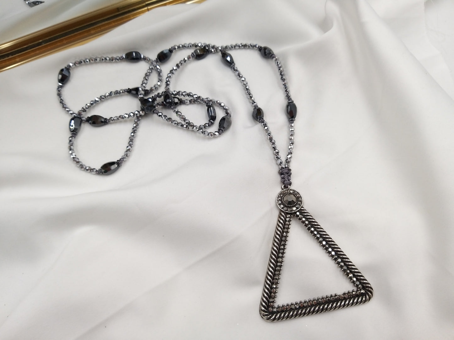 Κολιέ με αιματίτη και τριγωνικό μεταλλικό μενταγιόν πάνω σε λευκό ύφασμα – χειροποίητο κόσμημα euhandmade.