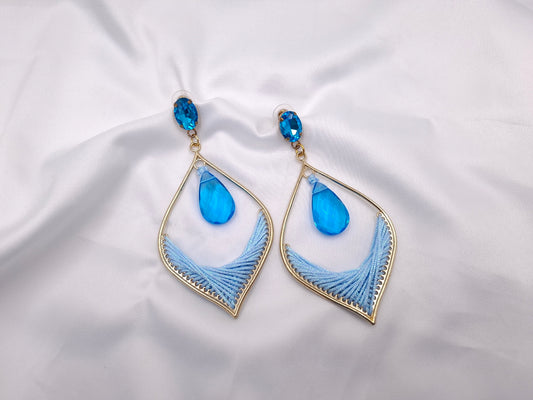 Χειροποίητα statement σκουλαρίκια euhandmade με γαλάζια κρυστάλλινη σταγόνα, χρυσαφί μεταλλικό πλαίσιο και πλεκτό σχέδιο σε λευκό φόντο.